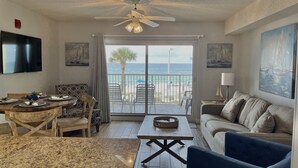 Un salon avec vue sur la plage, un ventilateur de plafond, un téléviseur et un coin repas avec une table mise pour deux personnes.