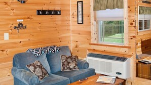 A cozy cabin interior with a blue sofa, a wooden coffee table, a wall-mounted heater, and a window with a view of greenery. - 1/2 double cabin with king bed and single child sized pull-out sleep sofa (sleeps 2 adults & 1 child (Flintstone)
