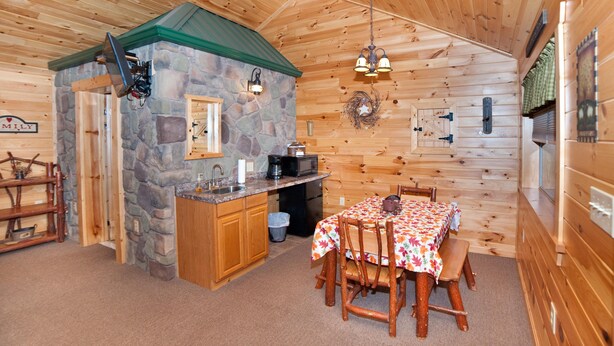 A cozy cabin interior with a stone fireplace, wooden dining table, and a kitchen area. - 1/2 double cabin with king bed and single child sized pull-out sleep sofa (sleeps 2 adults & 1 child (Flintstone)