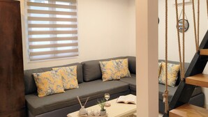 Una sala de estar con un sofá gris, cojines florales amarillos, una mesa de centro con velas y un libro, y una ventana con persianas.
