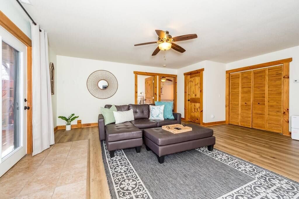 A living room with a sectional sofa, ottoman, patterned rug, ceiling fan, and wooden doors.