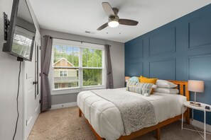Un dormitorio con cama, ventilador de techo, ventana con persianas y vista a una casa.