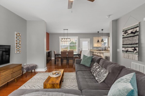 Una sala de estar moderna con un sofá gris, una mesa de centro de madera y un televisor. Hay un comedor con una mesa y sillas, y una cocina con armarios y una lámpara colgante.