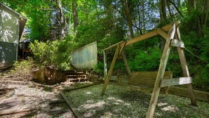 Legehus med gyngestativ i skovområde, grussti og træhegn.