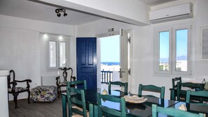 Una zona de menjador amb una taula, caires i una porta blava, amb vista al mar per la finestra.