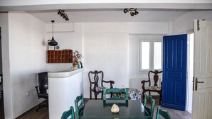 Una zona de menjador amb una taula de vidre, cadires verdes, una porta blava i una paret blanca amb una estant.