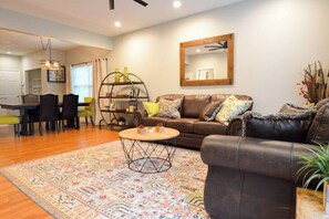 A living room with a brown leather sofa, a coffee table, and a patterned rug. There is a dining area with a table and chairs, and a kitchen visible in the background.