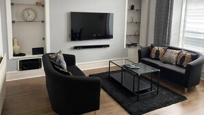 Una sala de estar moderna con un televisor de pantalla plana, sofás negros, una mesa de centro de vidrio y estanterías empotradas con objetos decorativos.