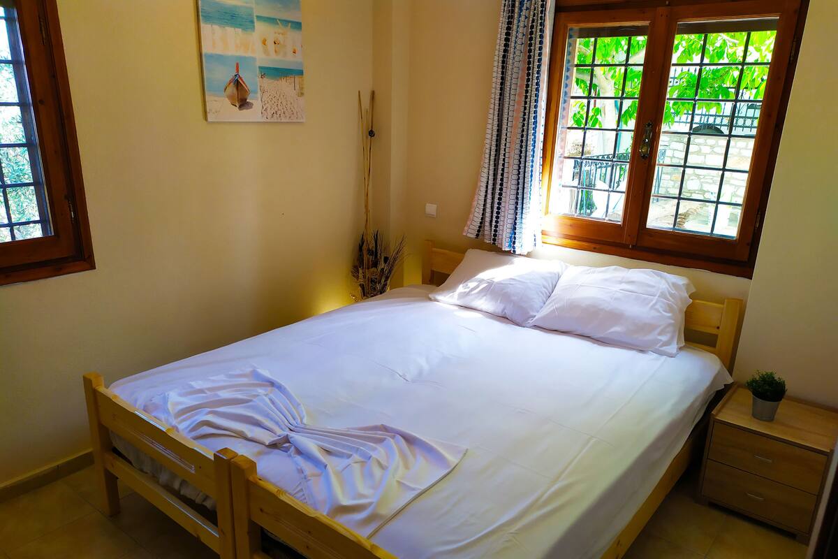 Una cama bien hecha con ropa blanca, un cabecero de madera, una mesita de noche con cajón, una pequeña planta en maceta y un cuadro de una playa en la pared.