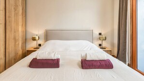 Una cama bien hecha con almohadas blancas y moradas, dos lámparas de noche y una ventana con cortinas.