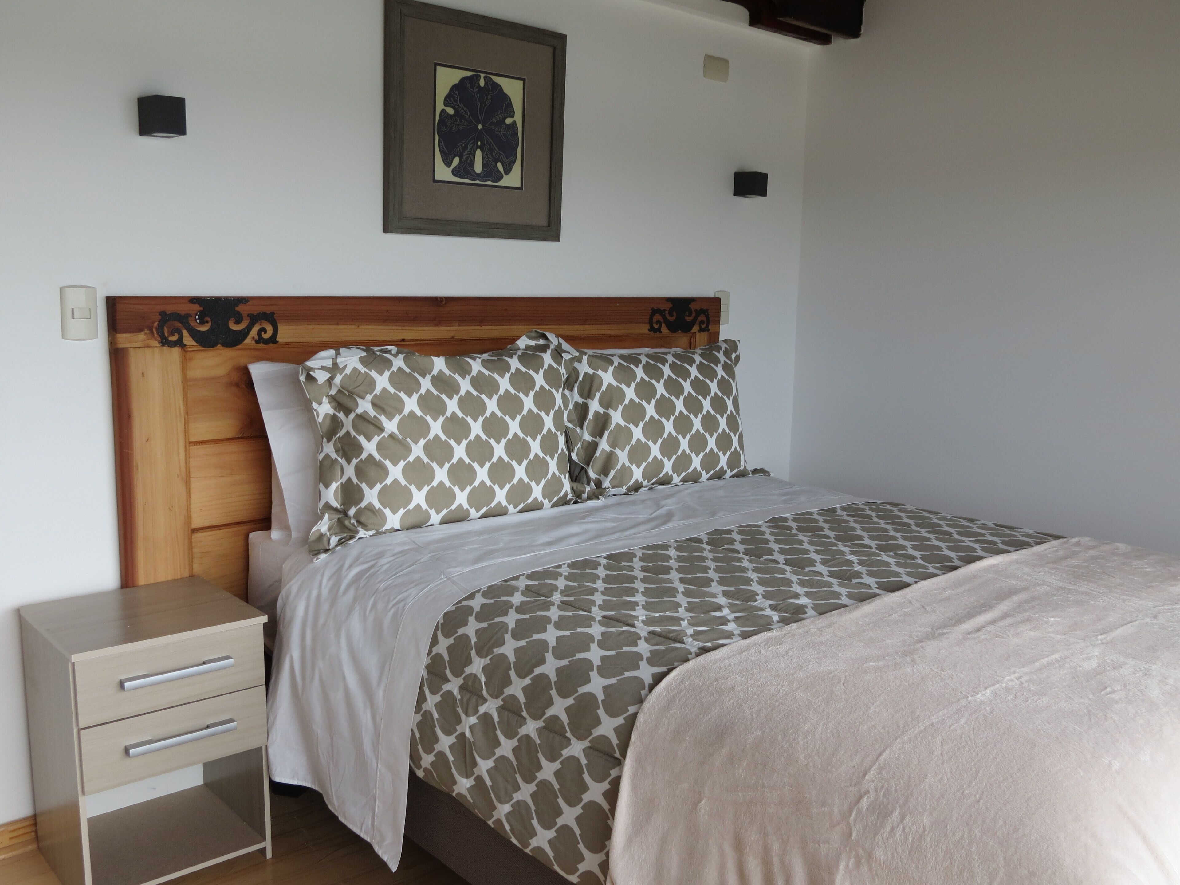 A neatly made bed with patterned bedding, a wooden headboard, a bedside table with drawers, and a wall-mounted light fixture.