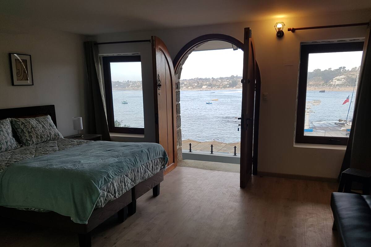 A bedroom with a bed, a window with a view of the sea, and a door leading to an outdoor balcony.