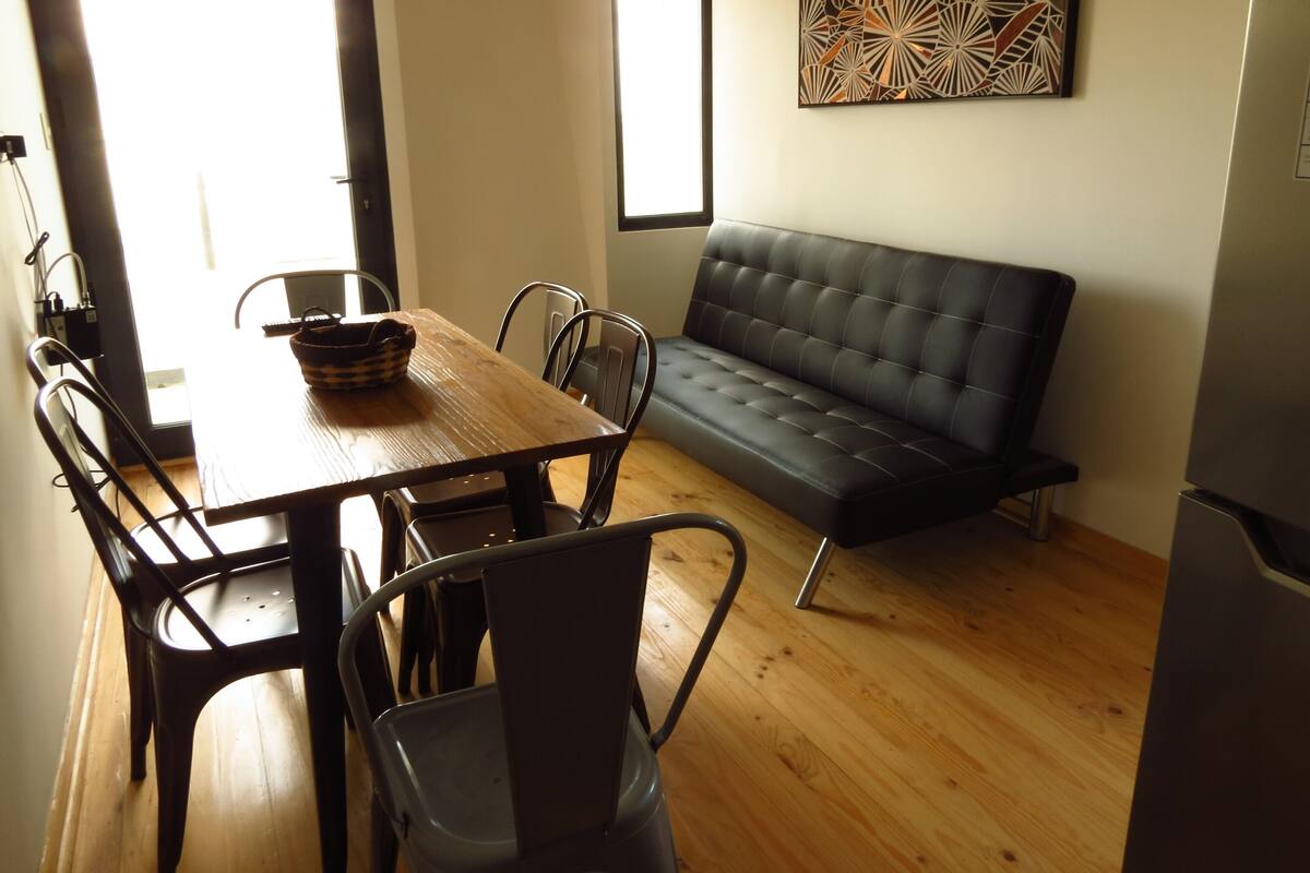 A dining area with a wooden table, metal chairs, a black sofa, and a wall-mounted abstract art piece.