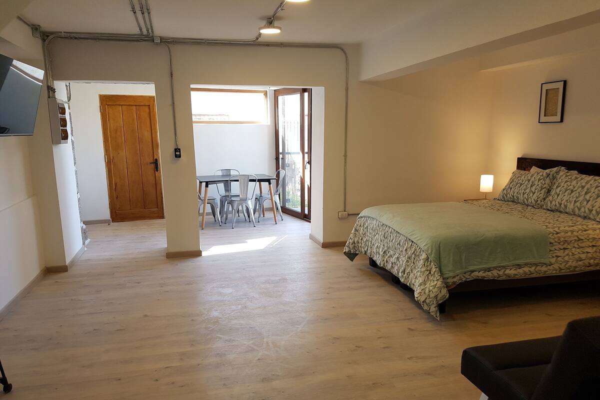 A spacious room with a large bed, a dining table with chairs, and a view through a glass door.