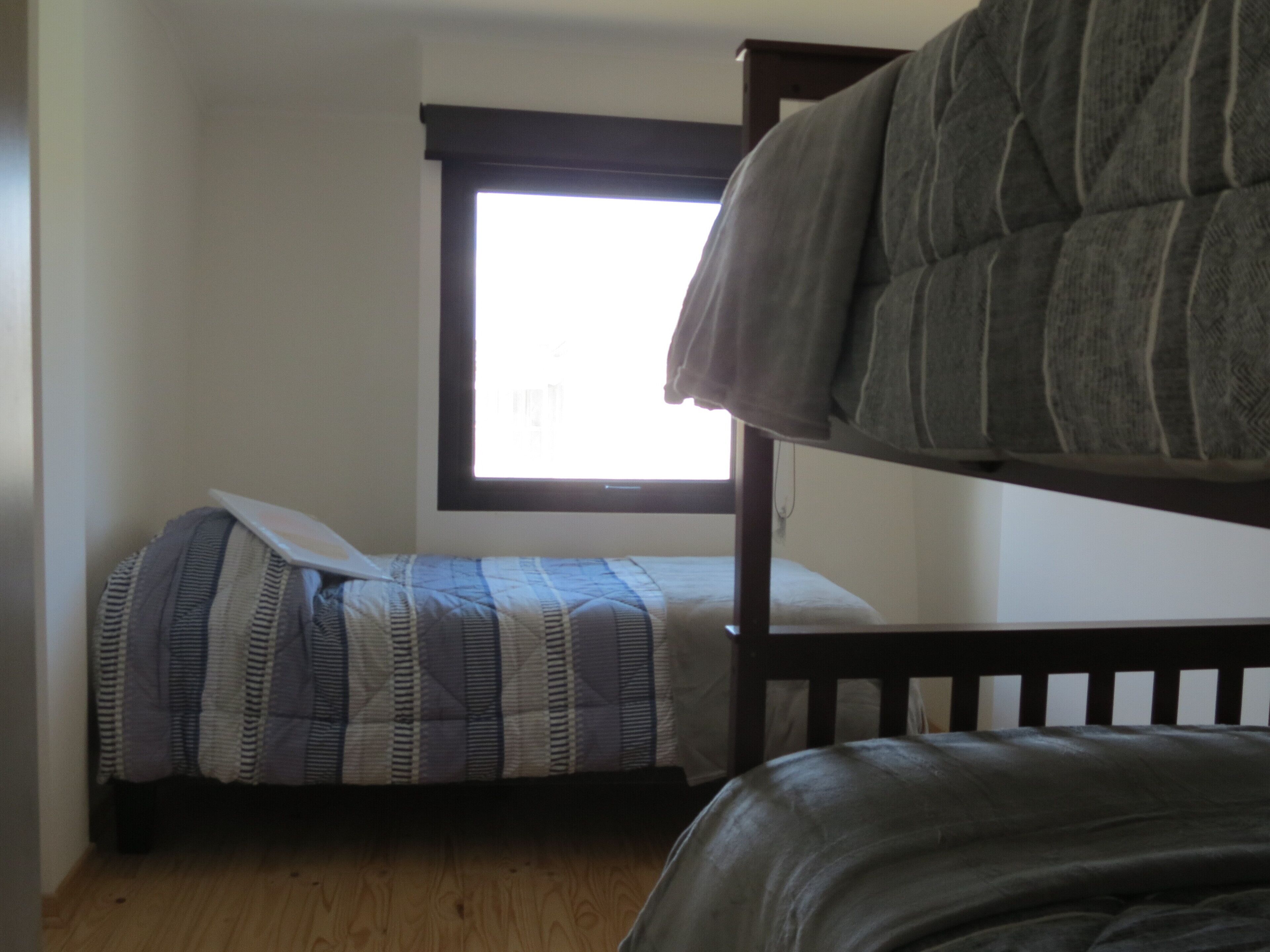 Una habitación con una litera y una cama individual, ambas con ropa de cama a rayas. Hay una ventana con persianas y el suelo es de madera.