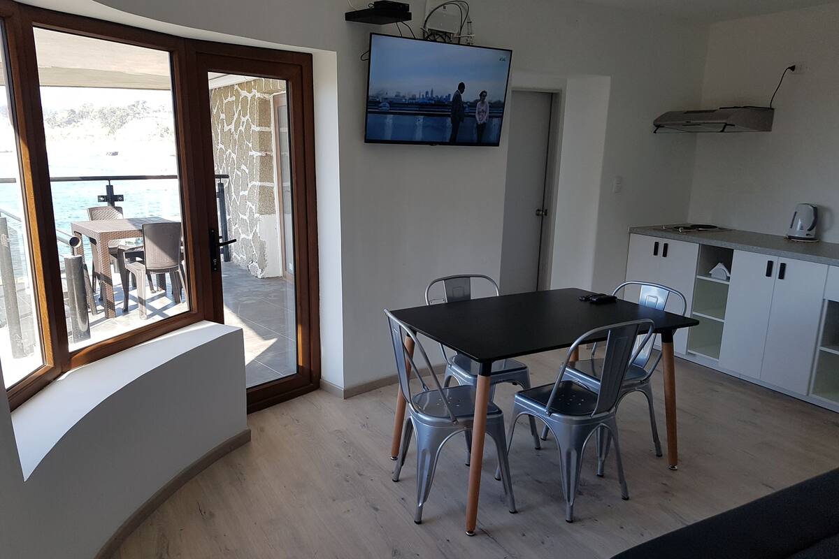 A modern kitchen with a dining area, a TV mounted above the counter, and a view of the sea through a large window.