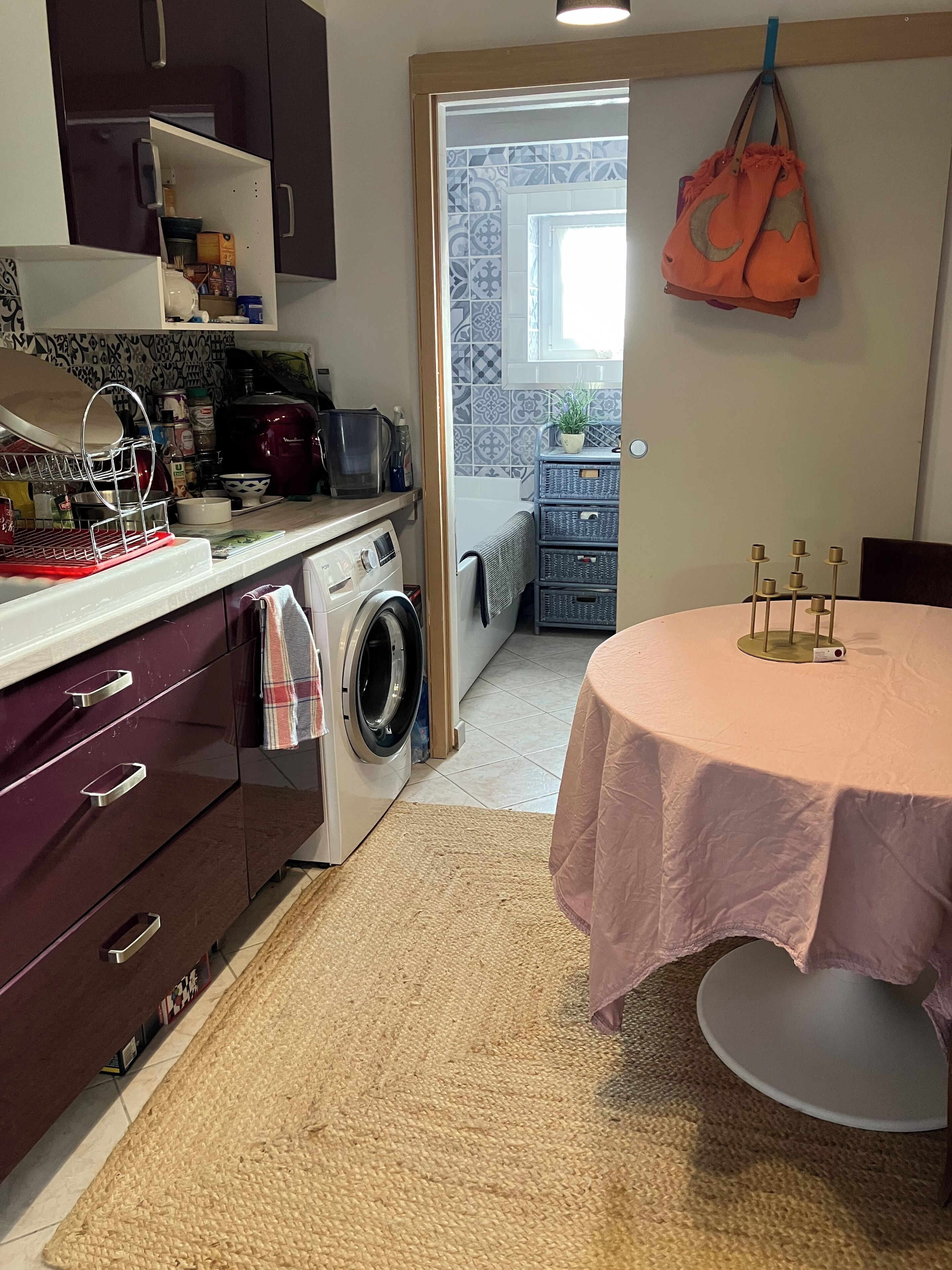 A compact kitchen with a washing machine, a microwave, and a sink. There is a dining table with a pink tablecloth and candle holders.