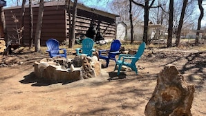 A rustic outdoor seating area with stone fire pits and blue chairs, surrounded by trees and a wooden structure in the background. - Charming cozy lake cabin, 300 yds from Lake Texoma beach, fishing, boat ramp! (Pottsboro)