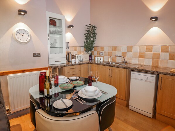 A modern kitchen with a dining table set for two, equipped with a dishwasher, a coffee maker, and a kettle.