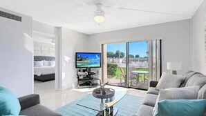 A modern living room with a flat-screen TV, a glass coffee table, and a sofa. There is a sliding glass door leading to a balcony with outdoor furniture. - Oceanfront walkout stunning condo! (Cape Canaveral)