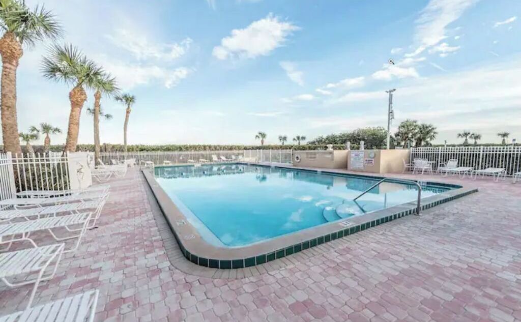 A rectangular swimming pool with a tiled edge, surrounded by a white fence and palm trees.