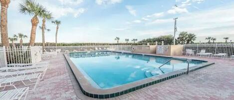 A rectangular swimming pool with a tiled edge, surrounded by a white fence and palm trees.