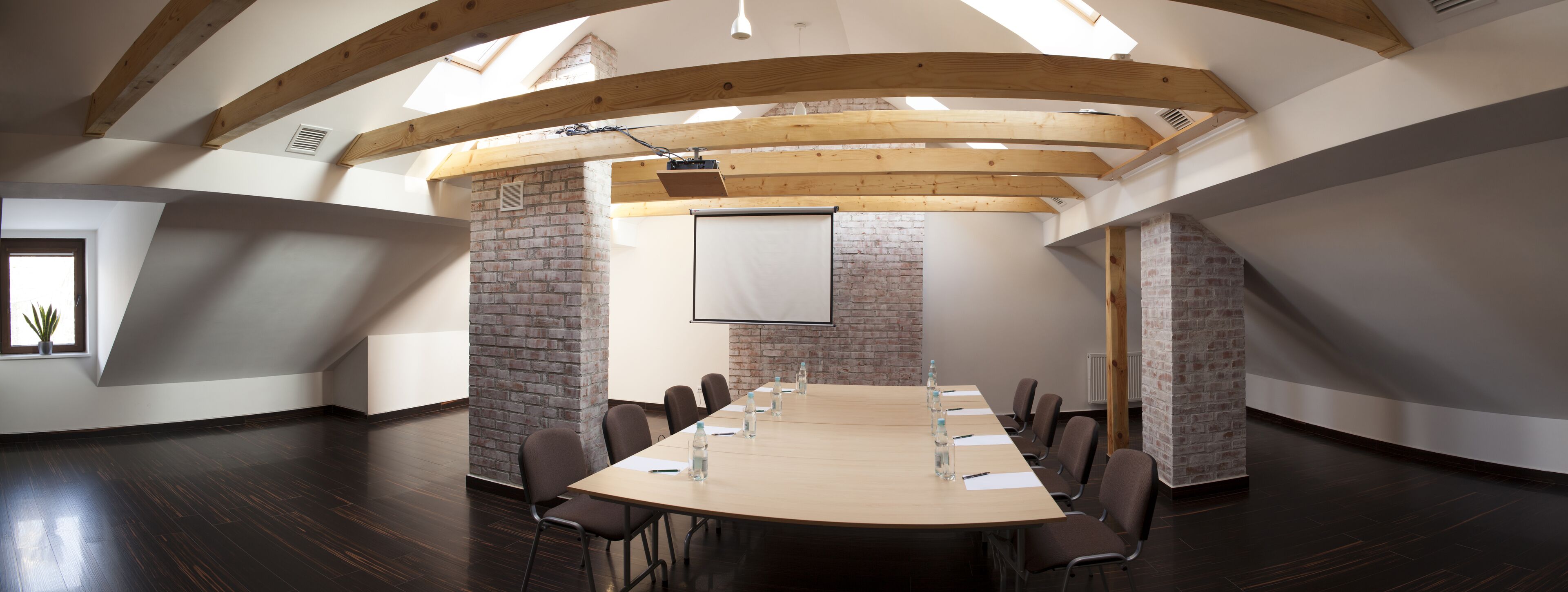 Un salón de conferencias con una larga mesa, sillas, una pantalla de proyector y paredes de ladrillo.