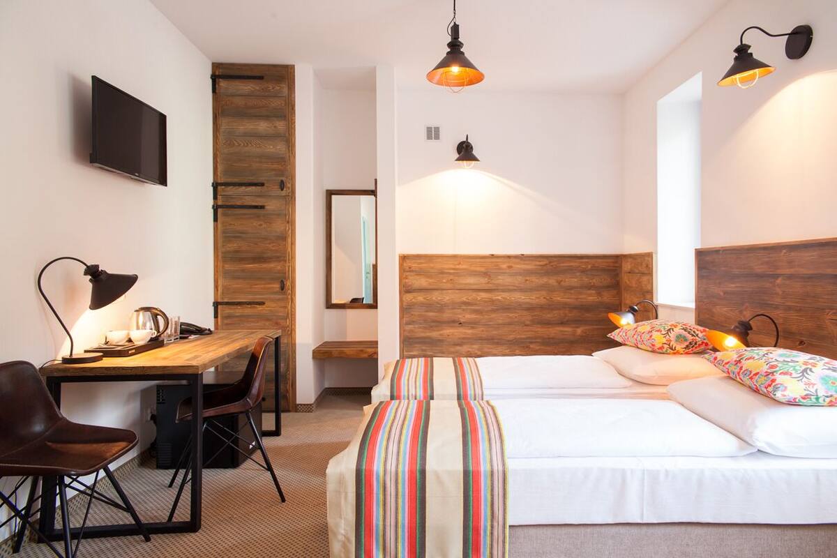 A hotel room with a wooden headboard, a bed with colorful pillows, a desk with a lamp, and a TV on the wall.