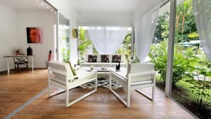 A modern living room with a white sofa set, a coffee table, and a dining table with chairs. There is a large window with sheer curtains, and a view of greenery outside. - Casa Blanca Villas Ojochal (Ciudad Cortés)