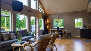 Ein geräumiges Wohnzimmer mit grauem Sofa, Korbstühlen, einem Esstisch und großen Fenstern mit Blick auf Grünflächen.