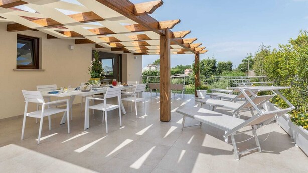 Une terrasse extérieure couverte, avec des chaises et des tables blanches, une pergola en bois et une vue sur la verdure.