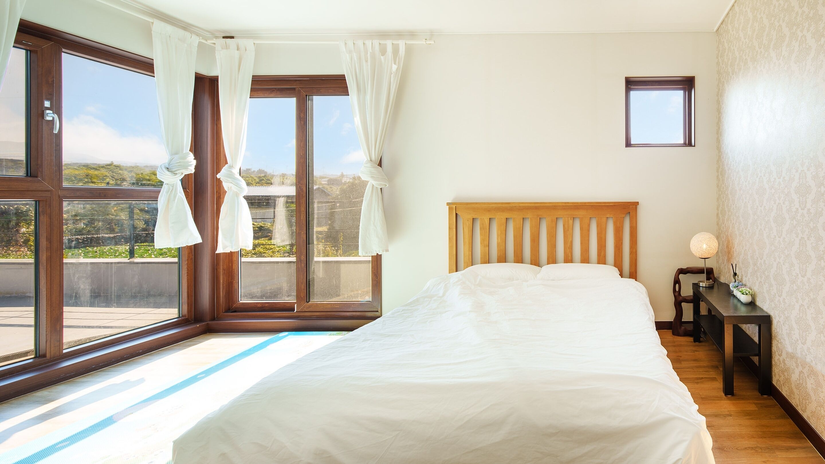 A bedroom with a large bed, wooden headboard, and a view of greenery through the windows.
