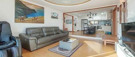 A living room with a sofa, a coffee table, and a TV. There is a painting on the wall and a kitchen in the background.