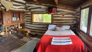 Ein gemütliches Blockhauszimmer mit einem Bett, einem kleinen Holztisch und einem Fenster mit Blick auf Bäume.