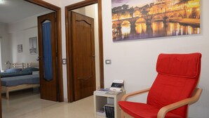 Una habitación con una cama, una silla roja, una mesita con revistas y un cuadro en la pared de un puente.