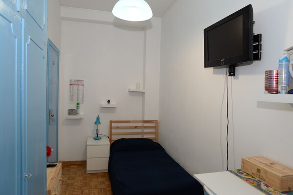 Una habitación pequeña y ordenada con una cama individual, una mesita de noche, un televisor montado en la pared y un armario de madera.