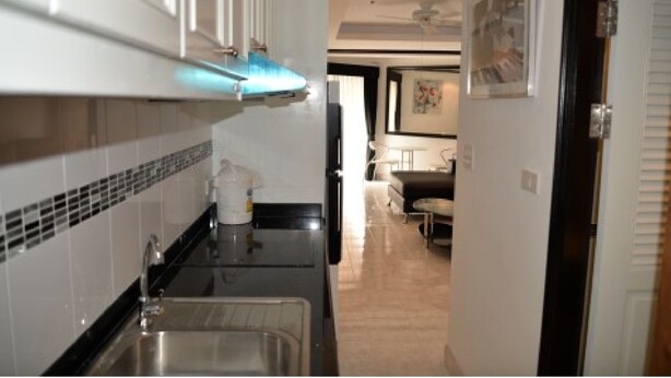 A narrow kitchen with a sink, cabinets, and a tiled backsplash. A dining area with a table and chairs is visible in the background.