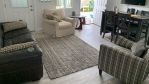 Una sala de estar con un sofá color crema, un sofá de cuero oscuro y un sillón a cuadros. Hay un televisor montado en la pared y una mesa de comedor con sillas.