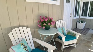 Un porche cubierto con dos sillas Adirondack blancas, una mesita redonda y una maceta con flores rosas.