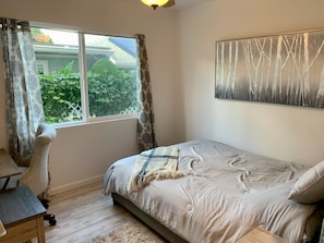 Un dormitorio con cama, escritorio y silla. Hay una ventana con cortinas, un cuadro en la pared y vistas al exterior con vegetación.