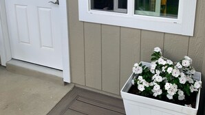 Una puerta blanca con un ventanal encima, un pequeño porche con suelo de madera y una jardinera blanca con flores blancas.