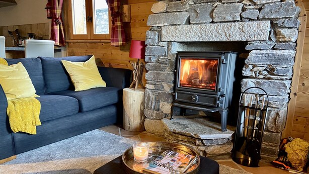A cozy living room with a stone fireplace, a blue sofa with yellow cushions, and a coffee table with magazines and a candle. - Sainte Foy Self-Catered Ski Apartment (Sainte Foy Tarentaise Station)
