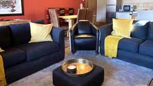 A living room with two dark blue sofas, a black leather chair, a small table with a candle, and a kitchen area in the background. - Sainte Foy Self-Catered Ski Apartment (Sainte Foy Tarentaise Station)