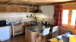 A kitchen with wooden cabinets, a microwave, a stove, and a sink. There is a bar counter with two chairs. The room has a wooden ceiling and a window with curtains. - Sainte Foy Self-Catered Ski Apartment (Sainte Foy Tarentaise Station)