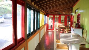 A covered outdoor seating area with a long table and chairs, red-framed windows, and a view of the street. - Pousada Saúde Melhor (Extrema)
