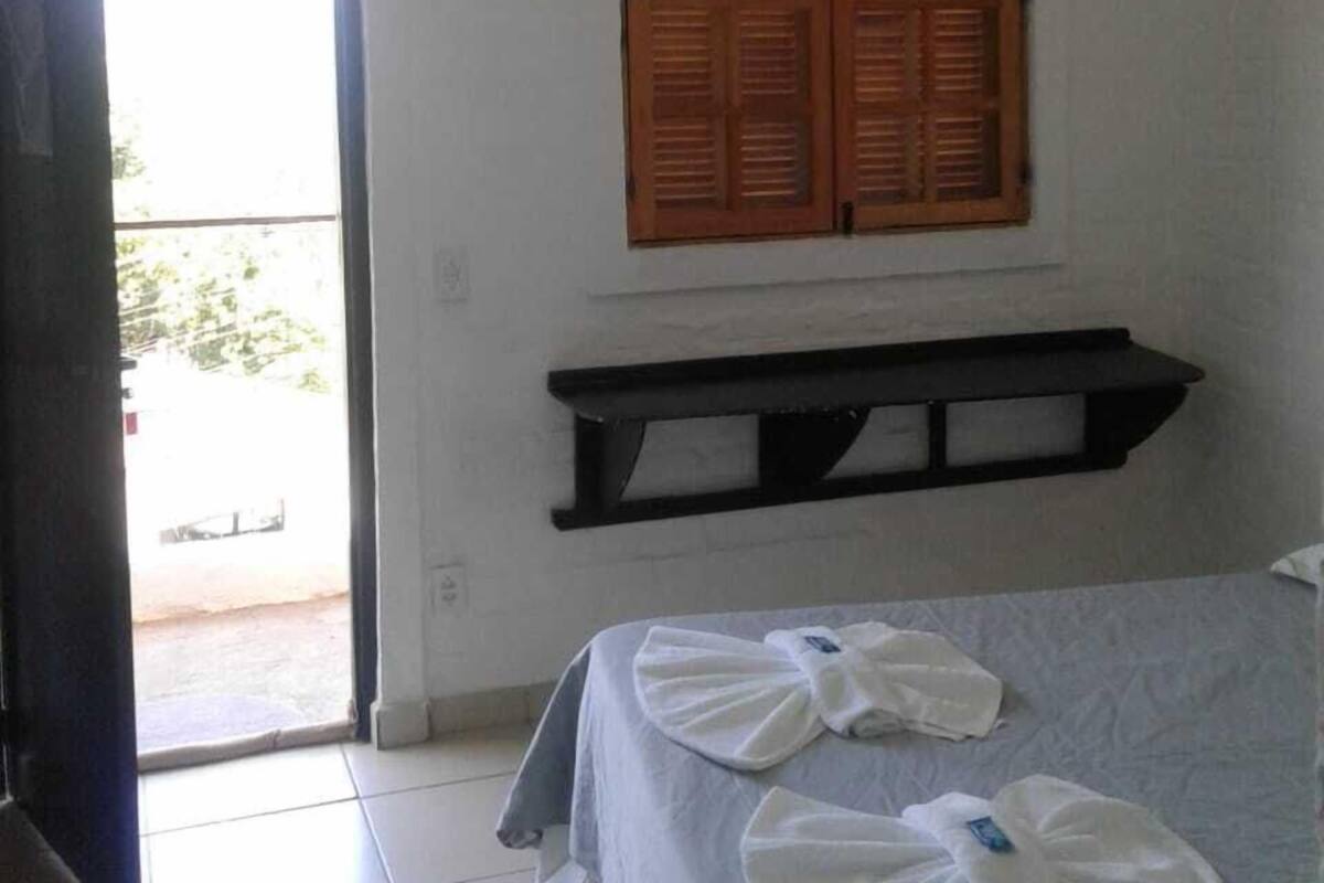 A hotel room with a bed, a wooden shuttered window, a black shelf, and a view of the outside.