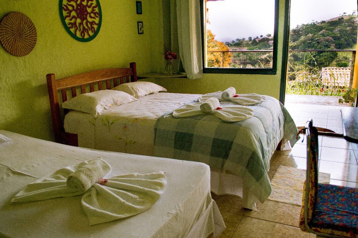 A hotel room with two beds, a wooden headboard, a window with a view of a hillside, and a small table with a chair.