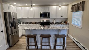 Een moderne keuken met witte kasten, roestvrijstalen apparatuur, een granieten aanrecht en drie houten krukken.