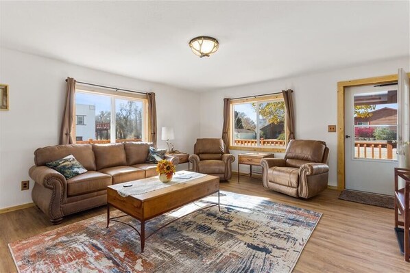 A spacious living room with a brown leather sofa, a wooden coffee table, and a large window offering a view of the outdoors. - Base Camp Cabin on Main (Hyattville)