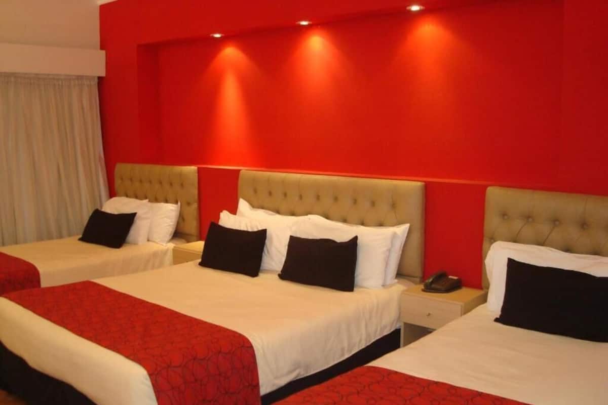 Camera d'albergo con tre letti, ognuno con copripiumino rosso e bianco, e una parete decorata in rosso.